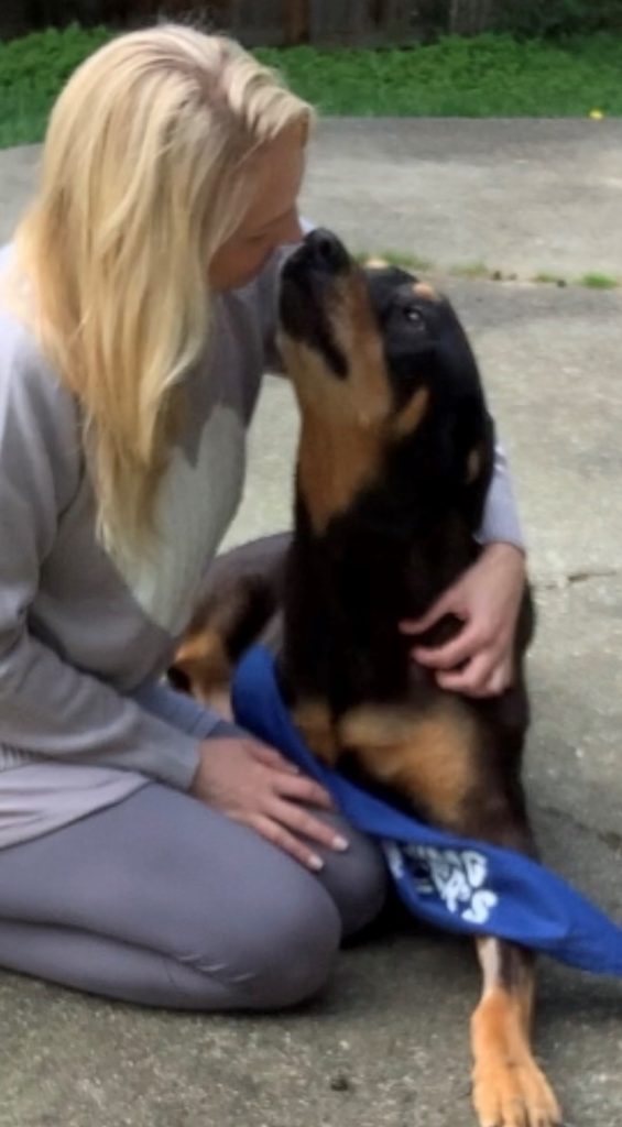 A blonde woman, Dr. Bethan Mullins, leans in for a kiss from a black and brown dog.